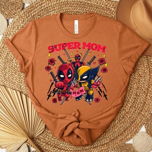 K&ouml;nnte beinhalten: Orangefarbenes T-Shirt mit einer Grafik von Deadpool und Wolverine und dem Text "SUPER MOM". Das Design beinhaltet Rosen und Schwerter. Das T-Shirt hat einen Rundhalsausschnitt und ein Etikett.