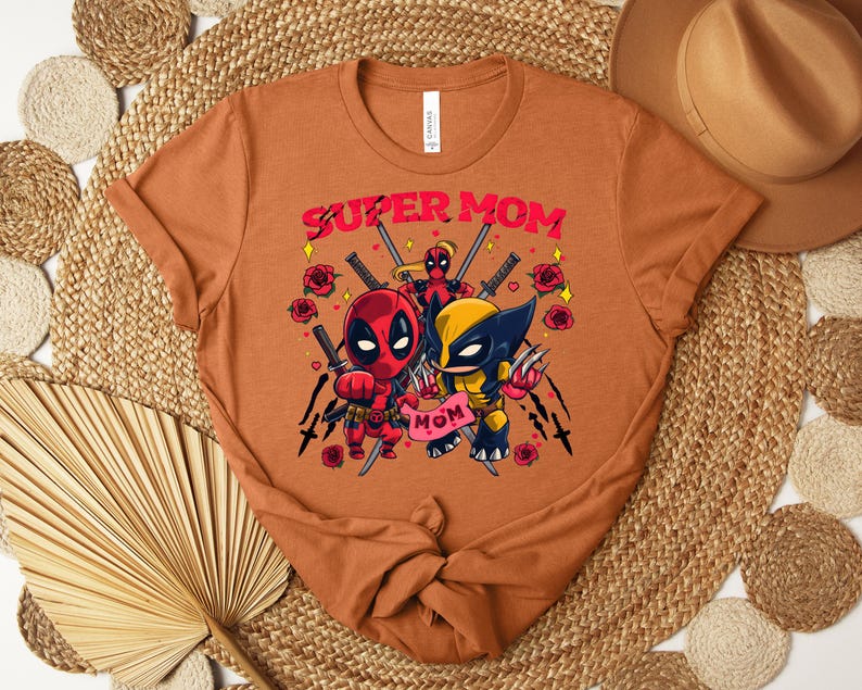K&ouml;nnte beinhalten: Orangefarbenes T-Shirt mit einer Grafik von Deadpool und Wolverine und dem Text "SUPER MOM". Das Design beinhaltet Rosen und Schwerter. Das T-Shirt hat einen Rundhalsausschnitt und ein Etikett.