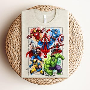 May include: Beige t-shirt with a vibrant illustration of superheroes such as Iron Man, Captain America, Spider-Man, Thor, Wolverine, Hulk, and Deadpool. The design is framed with a watercolour-style border. The shirt is folded on a woven surface.
