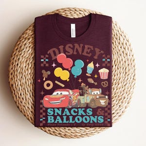 May include: A maroon t-shirt with a graphic design featuring the text "DISNEY" in a retro font, balloons, and cartoon cars. The shirt also includes the words "SNACKS & BALLOONS" and "Pixar Friends".
