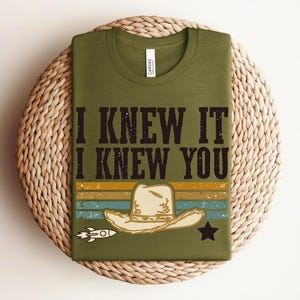 May include: An olive green t-shirt featuring the black text I KNEW IT I KNEW YOU above a graphic of a beige cowboy hat. The design includes horizontal stripes in yellow, blue, and orange, a small white rocket ship, and a black star icon.
