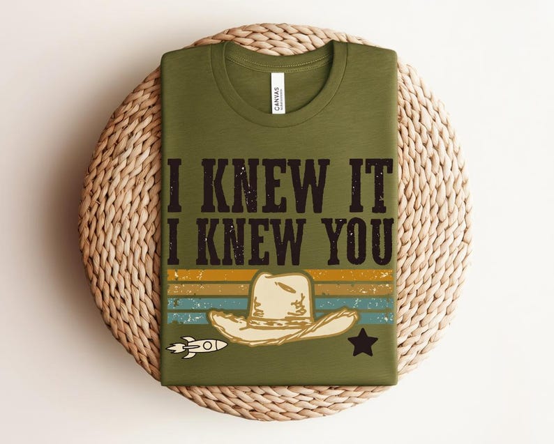 May include: An olive green t-shirt featuring the black text I KNEW IT I KNEW YOU above a graphic of a beige cowboy hat. The design includes horizontal stripes in yellow, blue, and orange, a small white rocket ship, and a black star icon.