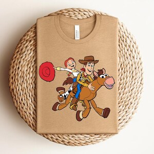 May include: A tan t-shirt featuring a colorful graphic of Woody, Jessie, and a horse from the Toy Story movie. The design shows the characters riding the horse. The shirt has a crew neck and short sleeves.