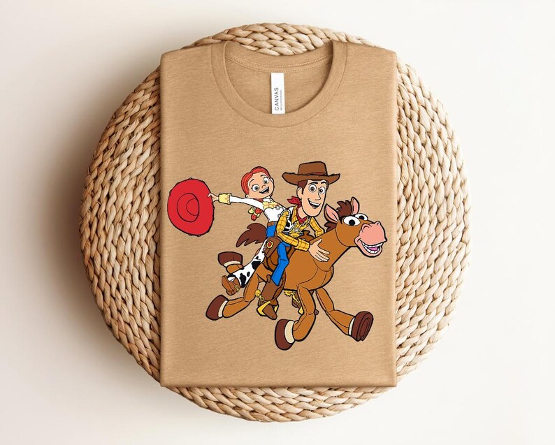 May include: A tan t-shirt featuring a colorful graphic of Woody, Jessie, and a horse from the Toy Story movie. The design shows the characters riding the horse. The shirt has a crew neck and short sleeves.
