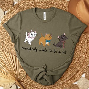 May include: Olive green t-shirt featuring three cartoon cats with the text "everybody wants to be a cat." The cats are white, tan, and black, each with unique accessories. The shirt is displayed on a woven surface.