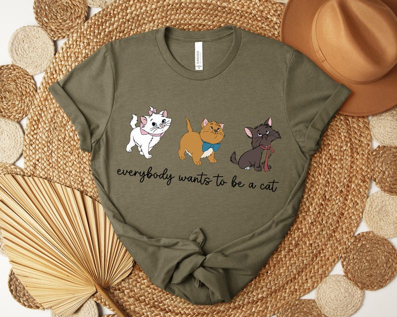 May include: Olive green t-shirt featuring three cartoon cats with the text "everybody wants to be a cat." The cats are white, tan, and black, each with unique accessories. The shirt is displayed on a woven surface.