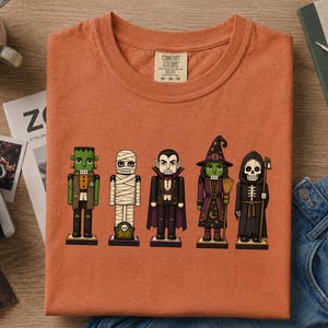 May include: A burnt orange t-shirt featuring a graphic print of five Halloween-themed nutcrackers. The characters include a green monster, a white mummy, a vampire in a cape, a green-skinned witch with a broom, and a black-robed skeleton.
