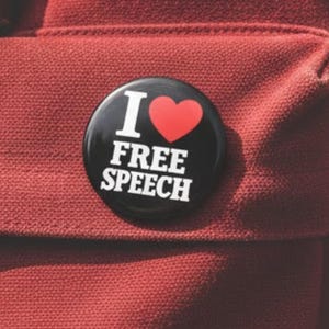 May include: A round, black button with the words "I ❤️ FREE SPEECH" in white text and a red heart. The button is pinned to a textured, reddish-brown fabric, possibly a backpack or bag.