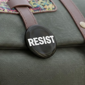 Puede incluir: Primer plano de un bolso de lona verde oliva con correas de cuero marrón y una solapa decorativa. Un botón ovalado negro con la palabra "RESIST" en letras blancas está sujeto al bolso.