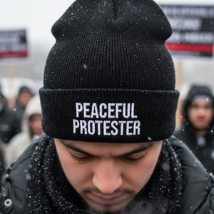 May include: A black knitted beanie hat with the words "PEACEFUL PROTESTER" in white. The hat is worn by a person, with other people and signs visible in the background. The image is taken outdoors, possibly during a demonstration.