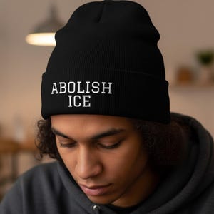 May include: Black beanie hat with the white text "ABOLISH ICE" embroidered on the front. The hat is made of a soft, knitted material. The person in the image is wearing a grey hoodie.