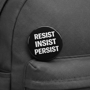 May include: A black button with the words "RESIST INSIST PERSIST" in white text is pinned to a black backpack. The button is round and the text is stacked vertically. The backpack is made of a textured fabric.