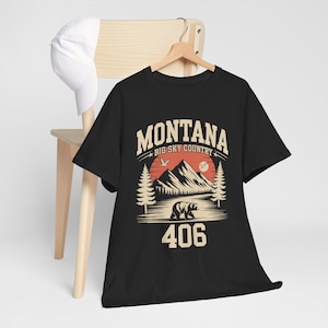 May include: Black t-shirt with a graphic design featuring the word "MONTANA" above a mountain scene with a bear and the number "406". The design includes a red sun and pine trees. A white baseball cap hangs on a wooden chair.