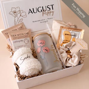 May include: A gift box labeled AUGUST Poppy containing two Tea With Tae packets, a frosted glass tumbler with a bamboo lid and poppy floral design, a pair of white fuzzy socks, a champagne-colored silk scrunchie, and gold under-eye masks.