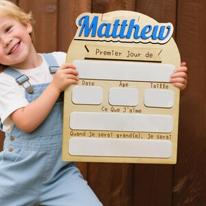 May include: A wooden sign with the name "Matthew" in blue script. The sign includes sections for "Date," "Age," and "Taille," along with the phrases "Premier Jour de," "Ce Que J'aime," and "Quand je serai grand(e), je serai."