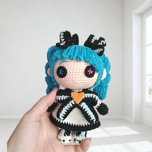 以下が含まれることがあります： 黒いリボンで結ばれた明るいティールブルーのツインテールの髪を持つかぎ針編みの人形。人形は淡いピンクの顔をしており、ピンクと青のステッチが施されたボタンの目があり、胸にオレンジ色のハートが付いた黒と白のドレスを着ています。