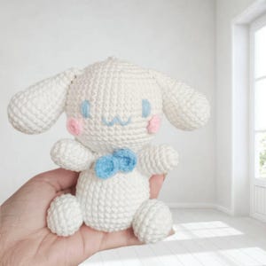 May include: White crocheted plush toy dog with blue and pink details. The dog has a blue bow tie and pink cheeks.