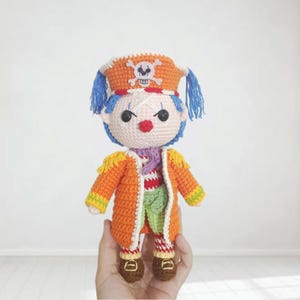 May include: A handmade crochet doll dressed as a pirate clown. The doll has blue hair, a red nose, and a hat with a skull and crossbones. It wears an orange coat with yellow trim, a striped shirt, green pants, and brown shoes.