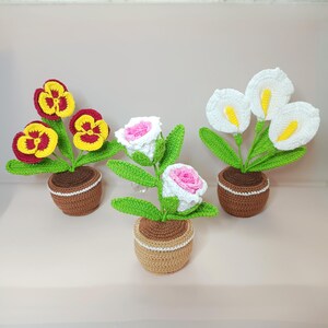以下が含まれることがあります： 3つの鉢植えの編み物花アレンジメント。最初のアレンジメントは、黄色と赤のパンジーが3つ。2番目のアレンジメントは、白とピンクのバラが2つ。3番目のアレンジメントは、白いカラーリリーが3つです。