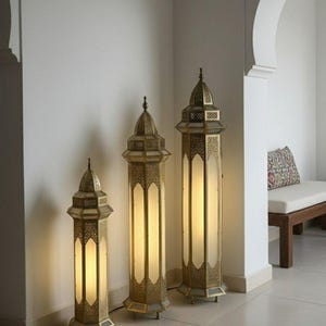 Può includere: Tre lampade da terra dorate con intricati disegni traforati e una luce calda. Le lampade, di altezze diverse, sono disposte contro un muro bianco. Le lampade sono in stile orientale.