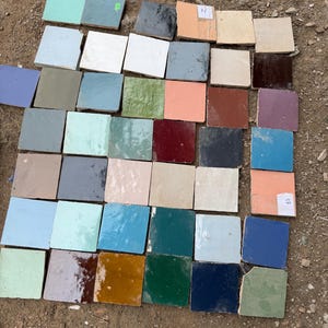 Può includere: Una collezione di piastrelle quadrate in ceramica di vari colori, tra cui blu, verde, bianco e marrone. Le piastrelle sono disposte su una superficie ruvida e terrosa, mostrando una gamma di trame e tonalità. Alcune piastrelle hanno etichette.
