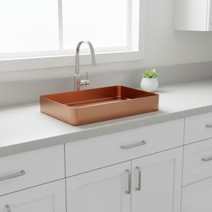 Könnte beinhalten: Ein rechteckiges Waschbecken in Kupferfarbe mit gebürsteter Oberfläche steht auf einer hellgrauen Arbeitsplatte. Ein silberner Wasserhahn befindet sich hinter dem Waschbecken. Das Waschbecken ist über weißen Schränken mit silbernen Griffen installiert.