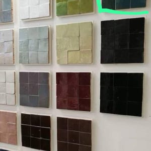 Pode incluir: Uma exposição de azulejos quadrados de cerâmica em várias cores, incluindo branco, verde, preto e tons de castanho e azul. Cada azulejo é dividido em nove quadrados menores, montados numa superfície branca. Os azulejos estão dispostos numa parede branca.