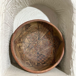 Peut inclure: Un bol rond en bois, orné de motifs géométriques complexes en marron et noir. L'intérieur du bol présente un design détaillé de triangles, de carrés et de lignes. Le bol est présenté dans une alcôve blanche.