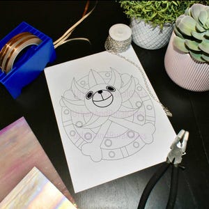 Puede incluir: Dibujo lineal en blanco y negro de una cara de oso de dibujos animados sobre papel blanco. También se ven un dispensador de cinta adhesiva azul, una cadena plateada y unos alicates.