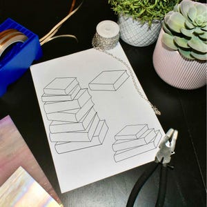 Puede incluir: Una hoja de papel blanca con un boceto a lápiz de libros apilados. Un recipiente azul con cinta de cobre, una cadena plateada y un par de alicates también son visibles sobre una superficie negra. Dos plantas en macetas están en el fondo.