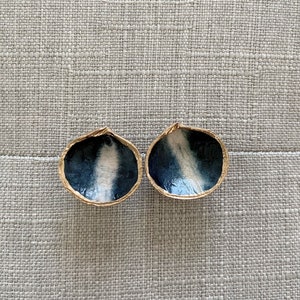 May include: Two halves of a walnut shell, each with a gold-painted rim and a dark blue and white interior design. The shells are displayed on a textured gray surface, creating a natural and artistic aesthetic.