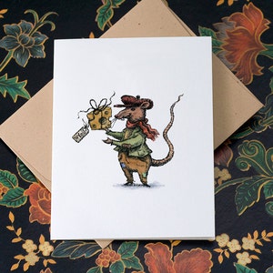 Puede incluir: Una tarjeta de felicitación blanca con una ilustración de una rata con boina, bufanda y chaqueta, sosteniendo un regalo de queso. La etiqueta del regalo dice "To You". La tarjeta va acompañada de un sobre marrón.