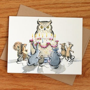 May include: A whimsical birthday card featuring an owl atop a cake with lit candles, surrounded by illustrated animals. The card is on a white background with a brown envelope, set against a wooden surface. The artwork has a watercolor style.