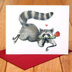 Könnte beinhalten: Aquarell-Grußkarte mit einem Waschbären, der eine rote Rose im Maul hält. Der Waschbär ist grau und weiß mit einem gestreiften Schwanz, und ein kleines rotes Herz schwebt darüber. Die Karte liegt auf einem roten Umschlag.