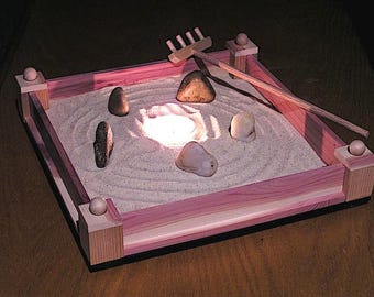 May include: A wooden zen garden with a sand tray, a small rake, and several stones. A small candle is lit in the center of the tray.