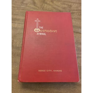 May include: A red hardcover book titled The Methodist Hymnal with a white cross and stylized M logo on the front cover. The text Dodge City, Kansas is printed in gold near the bottom. The book has a textured fabric cover and a vertical spine.