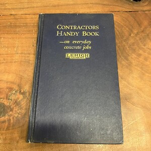 May include: A dark blue hardcover book titled Contractors Handy Book on everyday concrete jobs with the Lehigh Cement logo printed in gold on the front cover. The book features a rectangular shape with a textured fabric-like cover material.