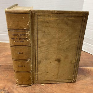May include: A tan hardcover book titled Annual Report of the Chief of Engineers U.S. Army 1907 Part 2. The book features a textured fabric cover with black horizontal lines on the spine and a thin black rectangular border on the front cover.