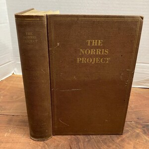 May include: A brown hardcover book titled The Norris Project with gold lettering on the front cover and spine. The book features a textured fabric cover with thin gold border lines and shows signs of wear on the edges and surface of the binding.