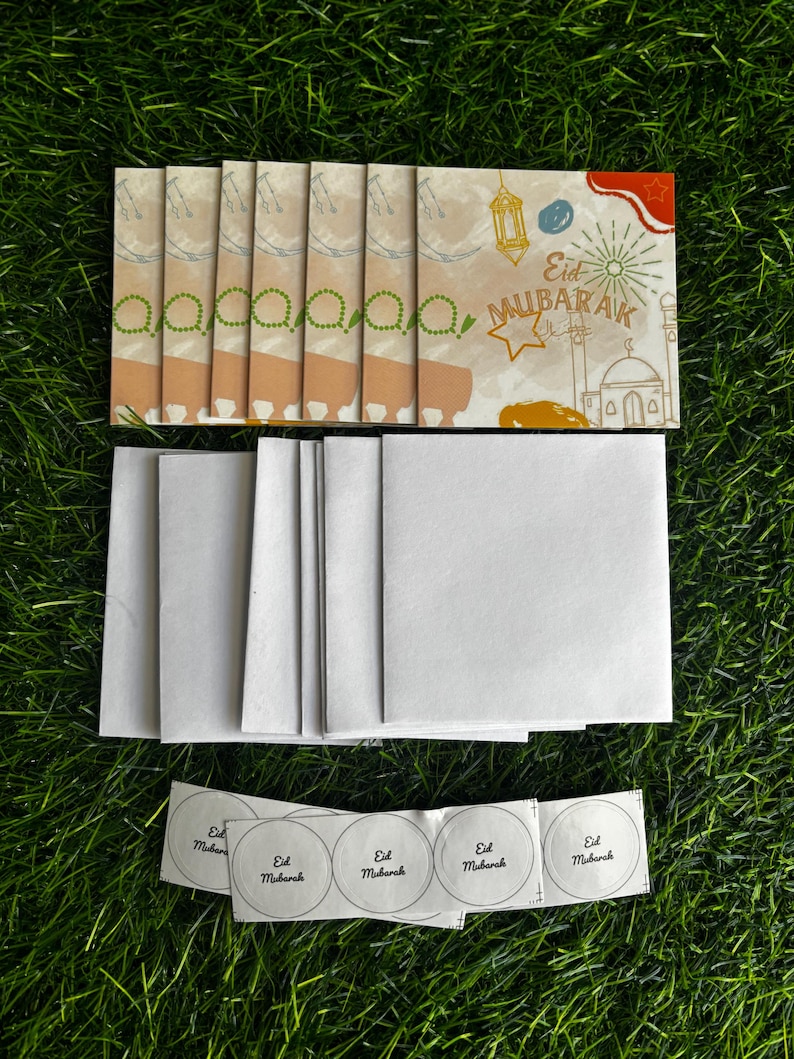 Puede incluir: Un conjunto de seis tarjetas de felicitaci&oacute;n en blanco con sobres y pegatinas. Las tarjetas son de color beige con un borde marr&oacute;n claro. Los sobres son blancos. Las pegatinas son redondas y blancas con el texto "Eid Mubarak" en negro. Las tarjetas presentan una ilustraci&oacute;n colorida de una mezquita, linternas y estrellas.