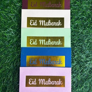 Puede incluir: Cinco sobres de diferentes colores con letras doradas en relieve que dicen "Eid Mubarak". Los sobres est&aacute;n apilados uno encima del otro sobre una superficie verde de c&eacute;sped.