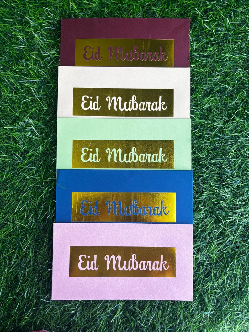 Puede incluir: Cinco sobres de diferentes colores con letras doradas en relieve que dicen "Eid Mubarak". Los sobres est&aacute;n apilados uno encima del otro sobre una superficie verde de c&eacute;sped.