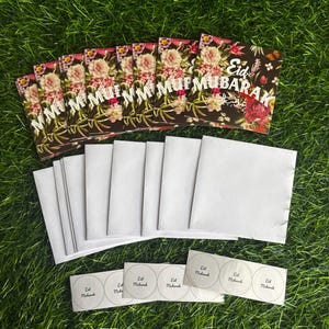 Puede incluir: Un conjunto de 10 tarjetas de felicitaci&oacute;n florales con el texto "Eid Mubarak" en letras blancas sobre fondo negro. Las tarjetas se acompa&ntilde;an de 10 sobres blancos y 5 pegatinas redondas con el texto "Eid Mubarak" en letras negras.