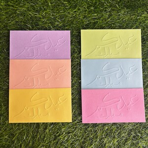 Puede incluir: Seis tarjetas de felicitaci&oacute;n con caligraf&iacute;a &aacute;rabe en relieve en varios colores, incluyendo rosa, naranja, amarillo y azul. Las tarjetas presentan la frase "Eid Mubarak" en &aacute;rabe.