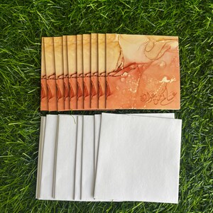 Puede incluir: Una pila de tarjetas de felicitaci&oacute;n en blanco con sobres blancos. Las tarjetas tienen un dise&ntilde;o de acuarela en tonos naranja, rojo y dorado. El texto de las tarjetas est&aacute; en &aacute;rabe.