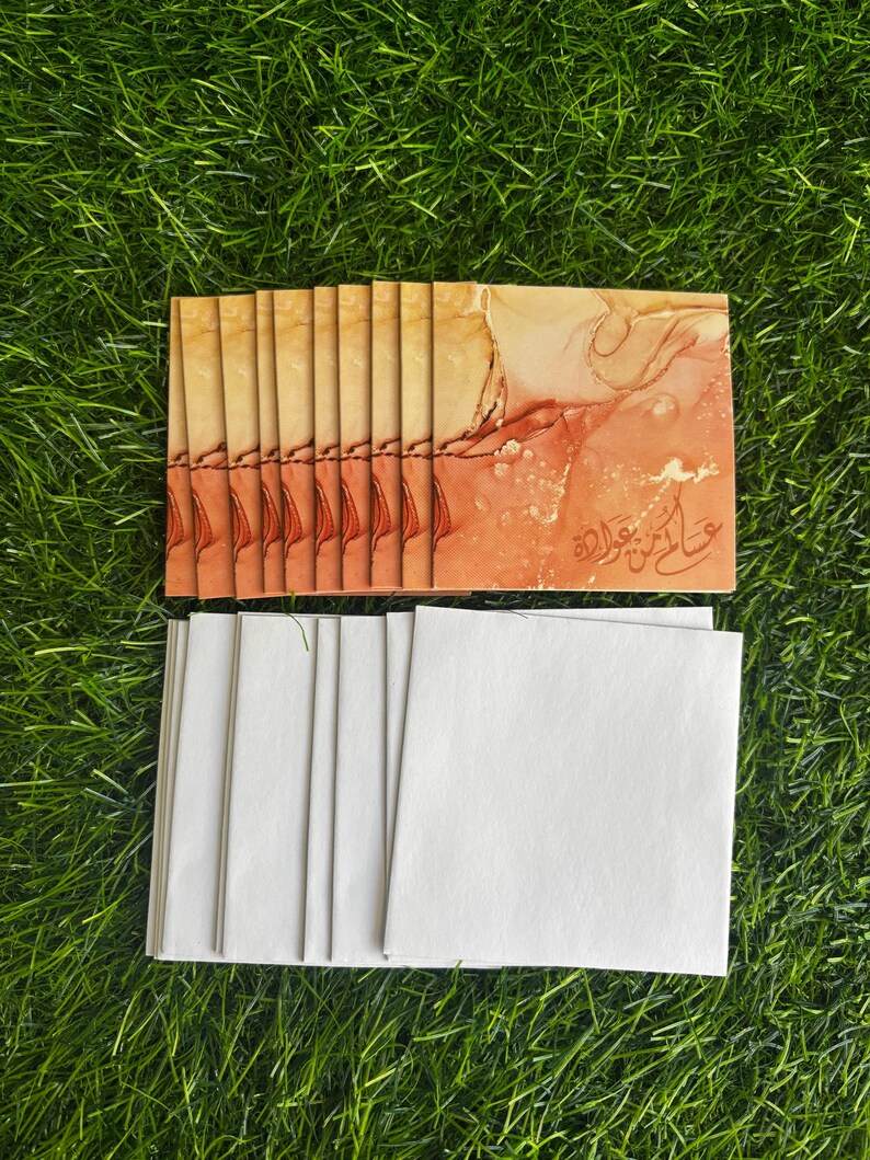 Puede incluir: Una pila de tarjetas de felicitaci&oacute;n en blanco con sobres blancos. Las tarjetas tienen un dise&ntilde;o de acuarela en tonos naranja, rojo y dorado. El texto de las tarjetas est&aacute; en &aacute;rabe.