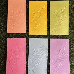 Puede incluir: Seis tarjetas de felicitaci&oacute;n en blanco con un dise&ntilde;o en relieve en escritura &aacute;rabe. Las tarjetas son de varios colores: naranja, amarillo, verde, rosa, azul claro y morado claro.