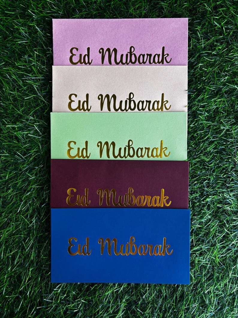 Puede incluir: Cinco sobres de diferentes colores con el texto "Eid Mubarak" escrito en letras doradas. Los sobres est&aacute;n apilados uno encima del otro, con el sobre rosa en la parte superior y el sobre azul en la parte inferior.