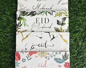May include: Four white greeting cards with floral designs and the text "Eid Mubarak" in different styles. The cards are stacked on top of each other on a green grassy surface.