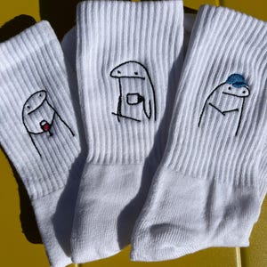 Könnte beinhalten: Drei Paar weiße gerippte Socken mit gestickten Cartoonfiguren. Jede Socke hat ein anderes Design: eine hält ein Getränk, eine mit einer blauen Kappe und eine mit einem roten Objekt. Die Socken werden auf einer gelben Oberfläche präsentiert.